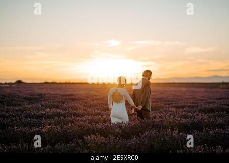 Sposo che tiene le mani dello sposo mentre cammina nel campo di lavanda sopra sfondo del cielo del tramonto il giorno del matrimonio Foto Stock