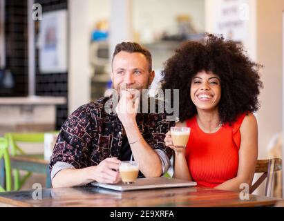 Una coppia multietnica soddisfatta, seduta al bar e trascorre del tempo insieme mentre ti godi il weekend e guarda la fotocamera Foto Stock