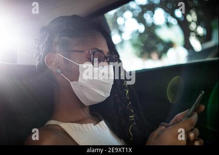 La giovane afrolatina in maschera facciale utilizza lo smartphone nel sedile posteriore di un'auto Foto Stock