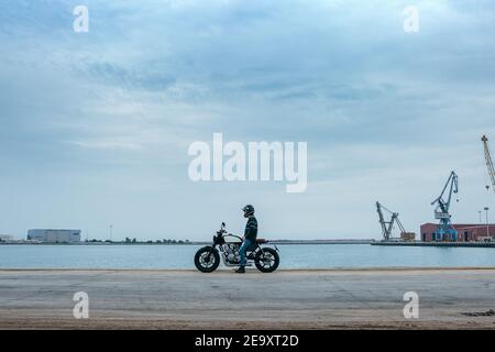 Vista laterale del biker maschile irriconoscibile in abiti casual e. casco di protezione seduto sulla motocicletta sul terrapieno vicino al mare a. cantiere Foto Stock