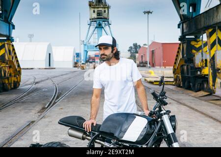 Vista laterale del biker maschile irriconoscibile in abiti casual e. casco di protezione seduto sulla motocicletta sul terrapieno vicino al mare a. cantiere Foto Stock