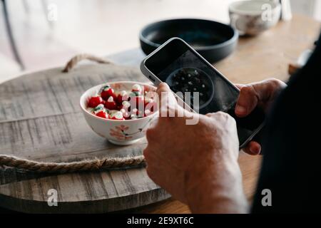 Vista dall'alto sul retro di uno chef maschile in uniforme irriconoscibile scattare foto sul cellulare del pasto in ciotola al ristorante Foto Stock