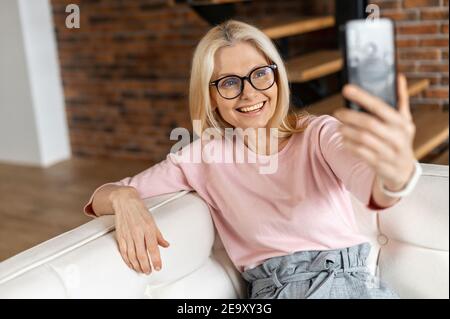 Sanguine allegra donna di mezza età prende un selfie sullo smartphone trascorrendo il tempo libero a casa. Matura affascinante donna indossare occhiali in posa a. Foto Stock