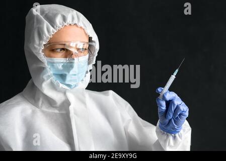 L'infermiere nei dispositivi di protezione individuale (PPE) contiene una siringa per l'iniezione del vaccino COVID-19. Ritratto di medico che indossa maschera e occhiali di protezione su una ba scura Foto Stock