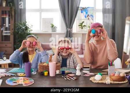 Ritratto di famiglia di tre seduti al tavolo e. tenendo le uova colorate davanti ai loro volti Foto Stock