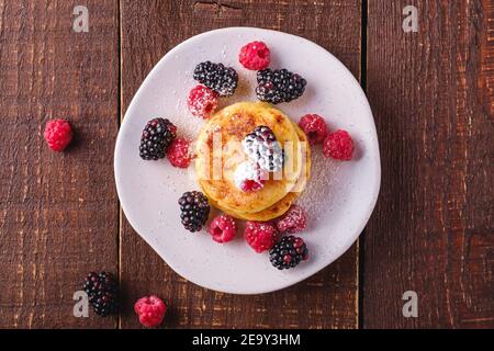 Frittelle di formaggio e zucchero in polvere, frittelle di cagliata dessert con lampone e frutti di bosco in piatto su sfondo di legno marrone scuro, top Foto Stock