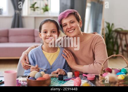Ritratto di madre felice abbracciando la figlia e sorridendo a. Macchina fotografica mentre dipingendo le uova di Pasqua al tavolo Foto Stock