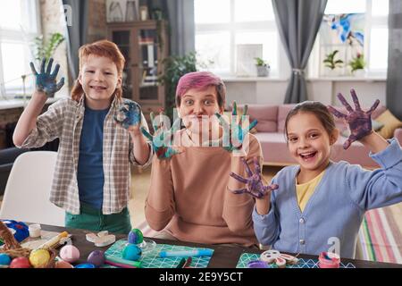 Ritratto di famiglia di tre con mani dipinte sorridenti Macchina fotografica mentre si prepara per Pasqua al tavolo Foto Stock