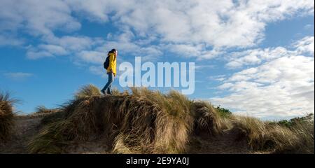 Donna escursionista che cammina lungo alte dune di sabbia. Giacca gialla e zaino contro un cielo blu. Foto Stock