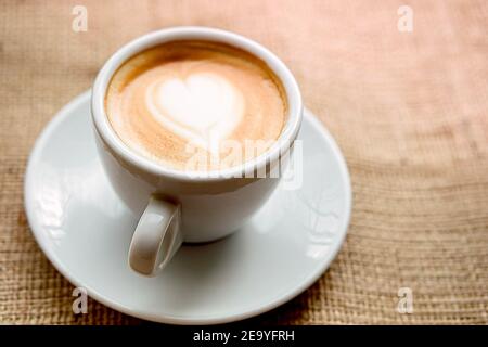 una tazza bianca su un piattino con una bevanda. caffè con latte, con cuore in schiuma si erge su un davanzale su tela di tela, solitudine Foto Stock