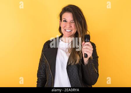 Giovane cantante ragazza, con giacca e microfono in pelle nera, verricello, su sfondo giallo. Foto Stock