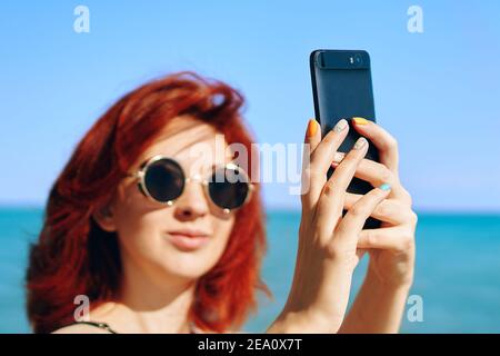 Selfie estivo. Bella donna scatta foto di se stessa sullo sfondo del mare blu e del cielo. Ragazza dai capelli rossi in occhiali da sole scuri prende selfie sulla fotocamera dello smartphone. Foto Stock