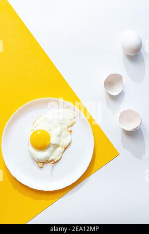 Uovo fritto su un piatto bianco, su fondo giallo e bianco. Composizione creativa minima con spazio di copia, vista dall'alto. Foto Stock