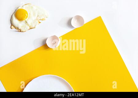 Uovo fritto su un piatto bianco, su fondo giallo e bianco. Composizione creativa minima con spazio di copia, vista dall'alto. Foto Stock