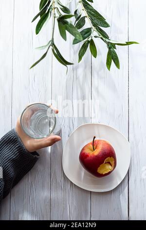 Concetto minimo di snack sano. Le mani del bambino tengono una mela e un bicchiere d'acqua. Ancora vita su un tavolo di legno chiaro con ombre e highli Foto Stock