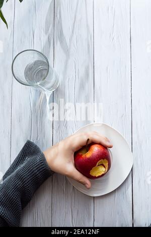 Concetto minimo di snack sano. Le mani del bambino tengono una mela e un bicchiere d'acqua. Ancora vita su un tavolo di legno chiaro con ombre e highli Foto Stock