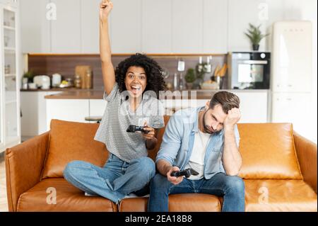 Una coppia multirazziale con i joystick si siede sul divano a casa e gioca al videogioco, una donna africana è vincitrice, gridare felice mentre il suo ragazzo Foto Stock