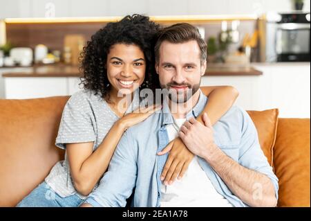Allegra coppia multietnica in amore si siede sul divano in abbraccia e guarda la fotocamera. Un ritratto teneramente di uomo sorridente e di ragazza africana che abbracciano Foto Stock