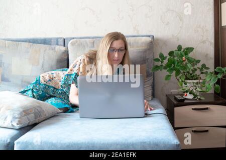 Donna europea con capelli lunghi bionda in occhiali si trova sul divano e guarda il monitor del computer portatile, isolamento in quarantena a casa, lavoro remoto Foto Stock