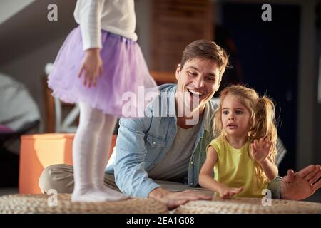 Piccole figlie che bevono sui mobili e ballano con il padre in un'atmosfera allegra a casa. Famiglia, casa, giocare, stare insieme Foto Stock