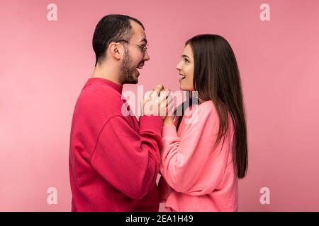 Foto di un uomo attraente con barba in abiti rossi e donna in rosa tiene per le mani l'una l'altra con la bocca aperta. Coppia sembra felice, isolato su rosa Foto Stock