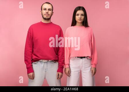 Foto di un uomo attraente con barba in abiti rossi e donna in piedi rosa con viso serio, isolato su sfondo rosa Foto Stock