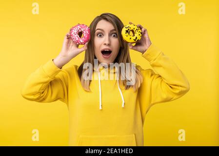 Foto di bella signora tiene le ciambelle davanti ai suoi occhi con bocca aperta e viso scioccato. Indossa felpa casual gialla con cappuccio, colore giallo isolato Foto Stock