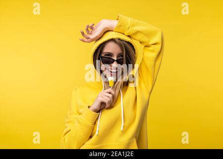 Foto di simpatica signora in bicchieri tiene lollipop e sorriso. Indossa un pullover casual giallo, isolato di colore giallo Foto Stock