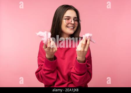 Foto di attraente signora in occhiali gioca con caramella di cotone, ha divertimento. Indossa casual rosa pullover bianco pantaloni isolato colore rosa sfondo. Foto Stock
