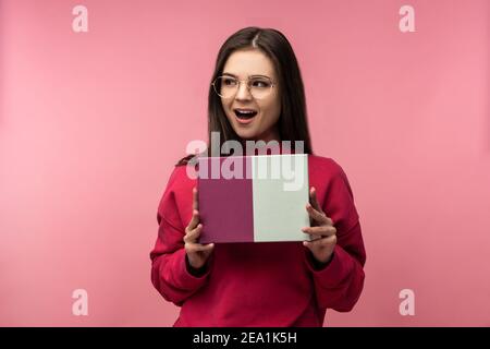 Foto di bella signora in bicchieri tiene scatola con regalo, attende e attende in attesa di aprire. Indossa pantaloni casual rosa pullover bianco colore rosa isolato Foto Stock