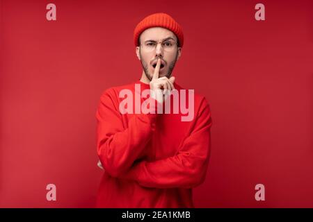 Foto di un uomo attraente con barba in occhiali e abiti rossi. Maschio scioccato e curioso, isolato su sfondo rosso Foto Stock