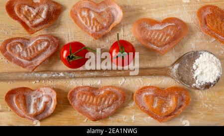 Ravioli freschi a forma di cuore con ripieno di pomodoro, cucchiaio di legno, pomodoro di ciliegia su un tagliere. Primo piano. Foto Stock