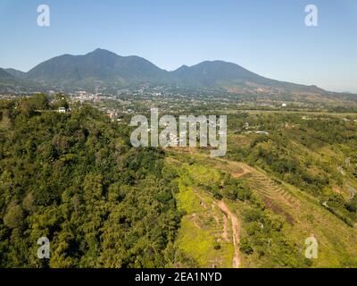 Vista aerea con droni di una montagna vulcanica con una piccola città e campi di riso alla sua base, circondata da una lussureggiante vegetazione tropicale a Bali, Indonesia. Foto Stock