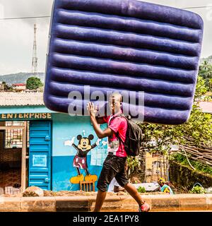 Un giovane trasporta un materasso ad aria gonfiata davanti ad un'azienda di Topolino nell'area rurale occidentale, Sierra Leone Foto Stock