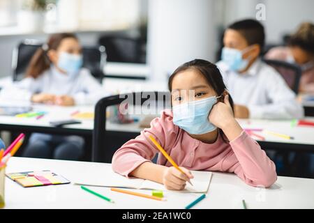 Ragazza della scuola asiatica in maschera seduta alla scrivania in classe Foto Stock