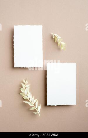 Schede di invito vuote. Disposizione piatta, vista dall'alto. Mockups bianchi vuoti di cancelleria di nozze con foglie essiccate su sfondo beige pastello. Foto Stock