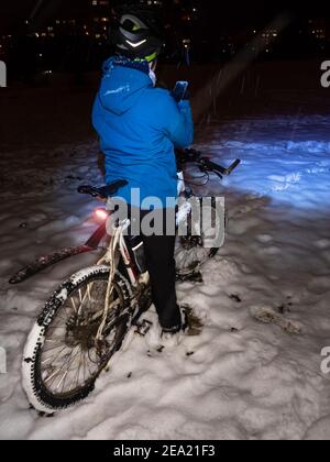 Vista posteriore dell'adolescente sedersi su mtb e scattare foto telefono di bicicletta nella neve. Il ragazzo indossa casco e faro luminoso. Serata invernale di nevica nel parco. Foto Stock
