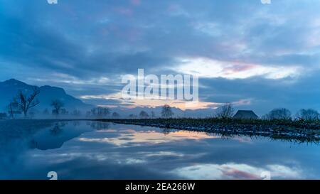 Tramonto con capanna, specchio d'albero, valle del Reno. Nuvole colorate e neve sul campo. Farbade Wolken beim Sonnenuntergang im Rheintal, Baum Spiegelung Foto Stock