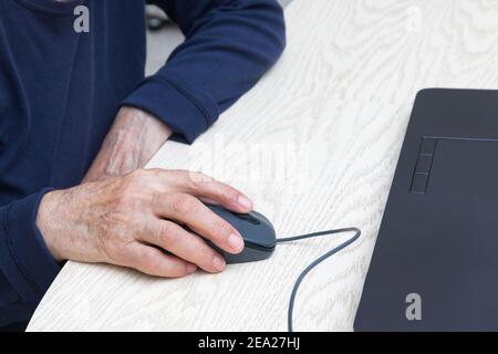 Mani di una donna anziana da vicino con un mouse per computer. Una donna in un pullover blu si siede a un tavolo e lavora su un computer portatile. Il concetto di riqualificazione o Foto Stock