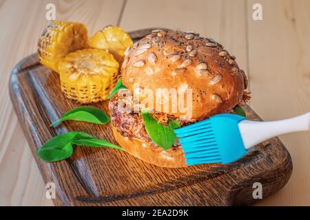 Uno chef o un fotografo usa un pennello per oliare un Burger su una tavola di legno prima di scattare una foto. Trucco e lifehack per rendere il pasto glisten e guardare delic Foto Stock