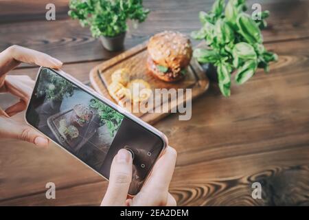 Girl food photographer and blogger scatta foto di un set di fast food con hamburger e patatine di mais su smartphone, quindi pubblica queste foto sulla rete sociale Foto Stock