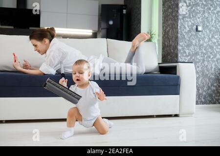 Il bambino gioca con la mop, mentre la madre naviga nel tuo smartphone sdraiato sul divano Foto Stock