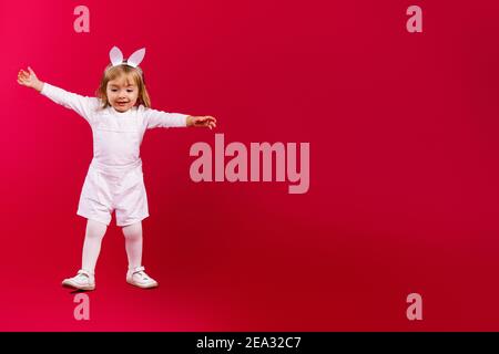 Foto a lunghezza intera di bella bambina che indossa orecchie conigliate di Pasqua e costume bianco, saltando su sfondo rosso con ampio spazio laterale per il testo: Vendita Foto Stock