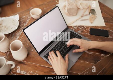 Primo piano ad alto angolo della digitazione delle mani maschili sul computer portatile con schermo bianco vuoto alla scrivania in officina, concetto di piccola impresa, spazio per la copia Foto Stock