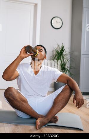 L'uomo afroamericano si rilassa sul tappeto da ginnastica dopo l'allenamento e beve una bottiglia di acqua dolce. Foto Stock