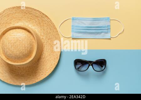 Ampio cappello di paglia di brim, occhiali da sole e maschera medica viso su sfondo giallo blu. Protezione contro il covidio e l'influenza durante le vacanze estive, i viaggi. Foto Stock