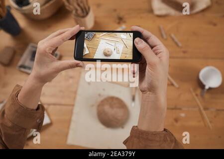 Primo piano ad angolo di una giovane donna che scatta foto smartphone di ciotola di ceramica grezza in laboratorio di ceramica, concetto di piccola impresa, spazio di copia Foto Stock