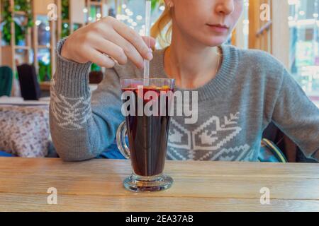 Una giovane donna in un maglione grigio si siede a un tavolo in un caffè di fronte a un bicchiere di VIN brulé. Messa a fuoco selettiva. Foto Stock