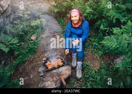 Buona donna che cucina gustoso barbecue e pollo kebab su una griglia. Lei usa il fuoco di campo in foresta. Concetto di picnic esterno Foto Stock