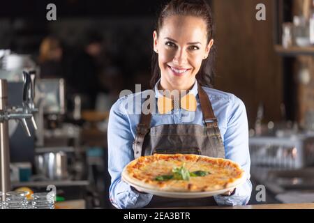 La cameriera sorride mentre serve una grande pizza italiana autentica in un ristorante. Foto Stock